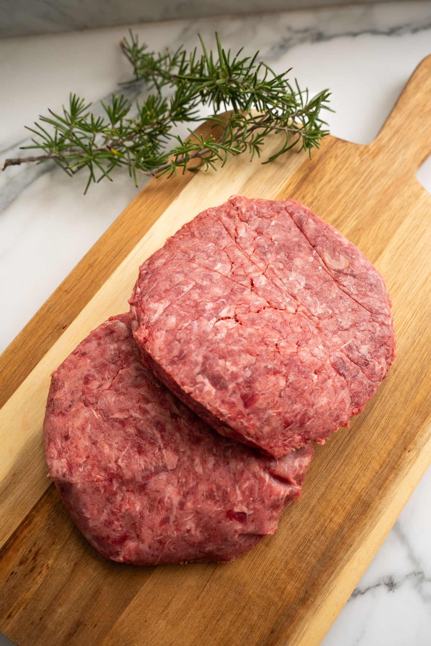 Two raw hamburger patties, resting ontop of a wooden butcher block with rosemary in the background
