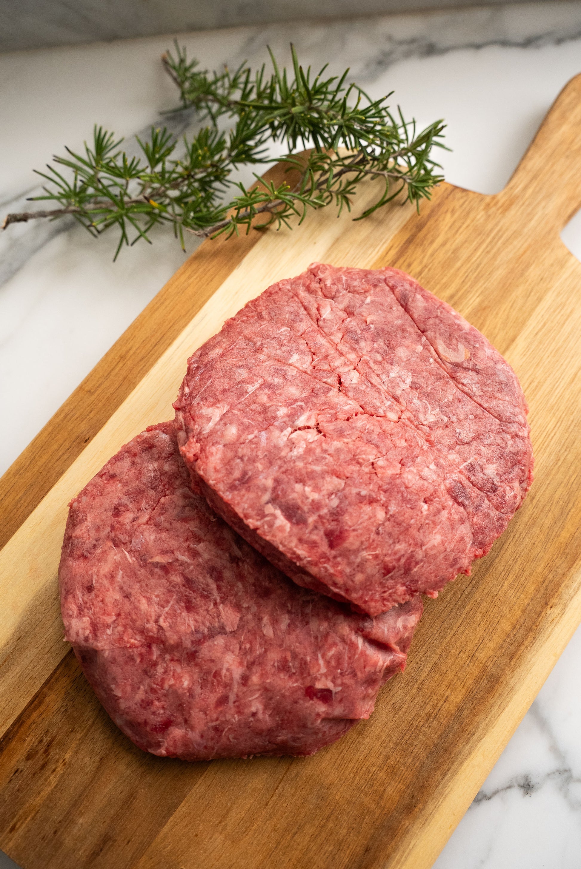 Two raw hamburger patties, resting ontop of a wooden butcher block with rosemary in the background