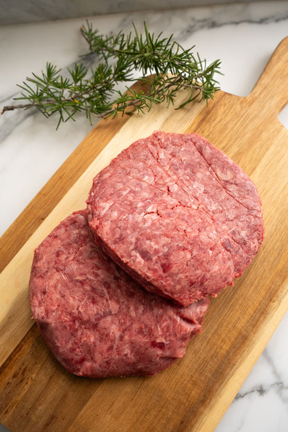 Two raw hamburger patties, resting ontop of a wooden butcher block with rosemary in the background