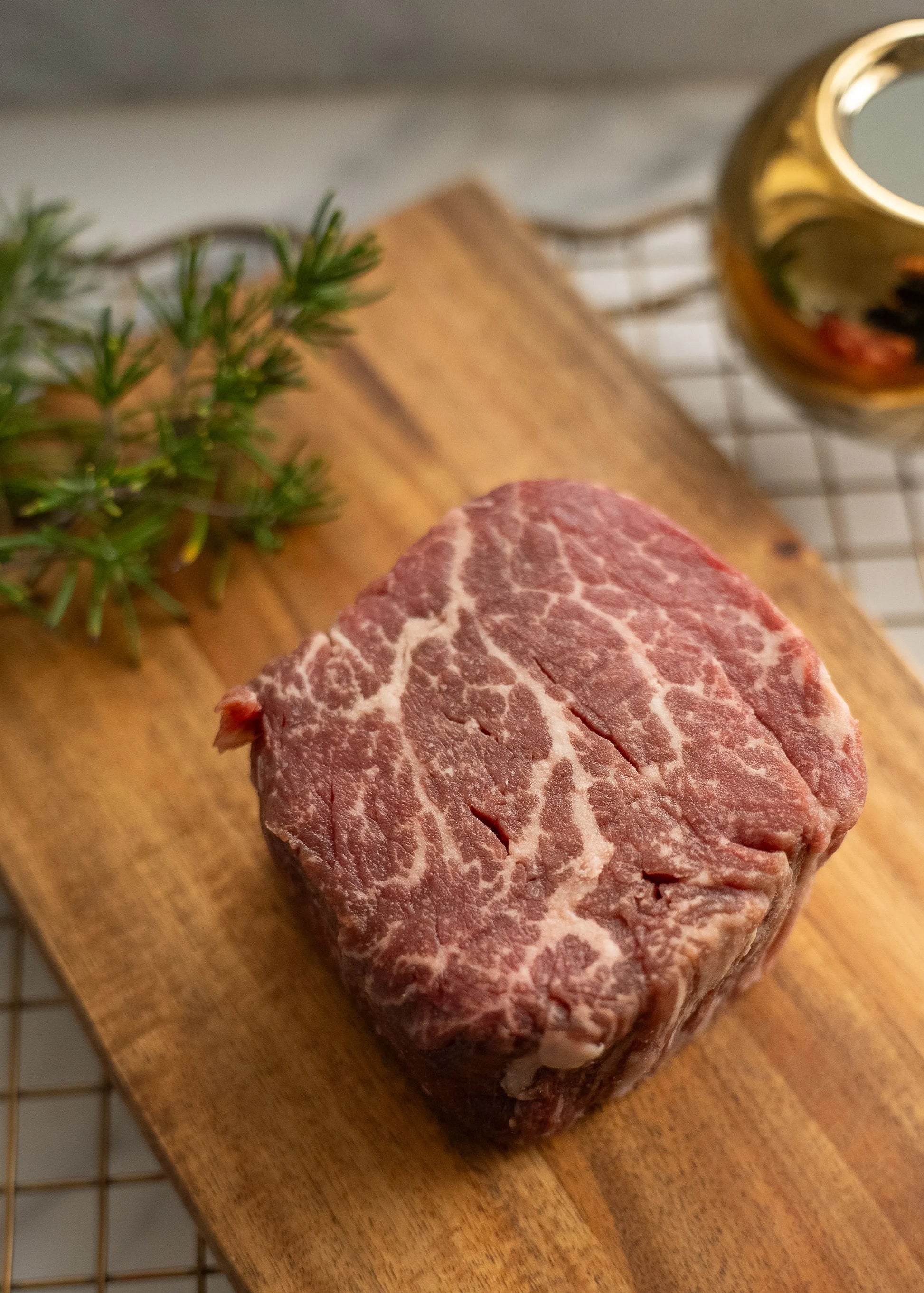 Raw Halal Wagyu  steak on a wooden cutting board with rosemary and a glass of red wine in the background.

