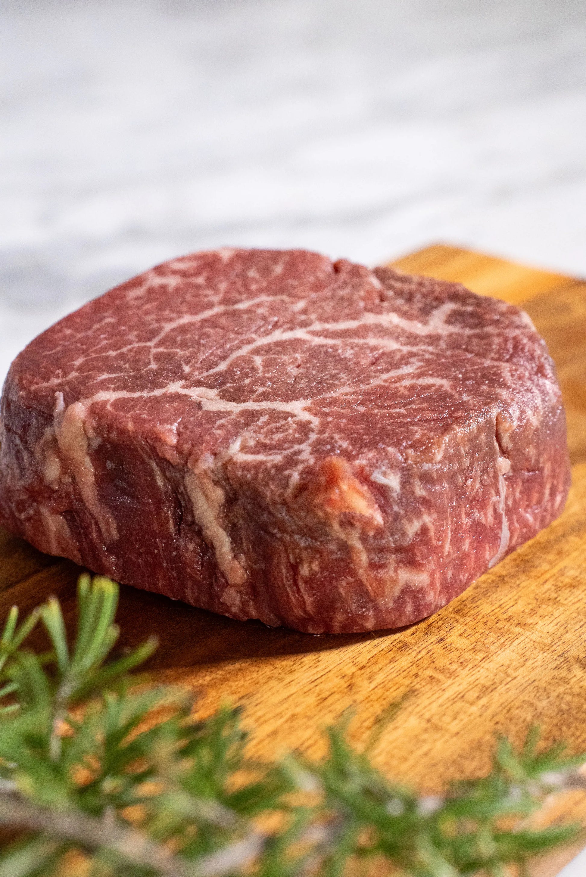 Raw Halal Wagyu filet steak on a wooden cutting board with rosemary on a a white granite top.

