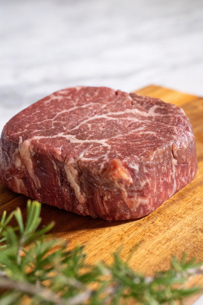 Raw Halal Wagyu filet steak on a wooden cutting board with rosemary on a a white granite top.

