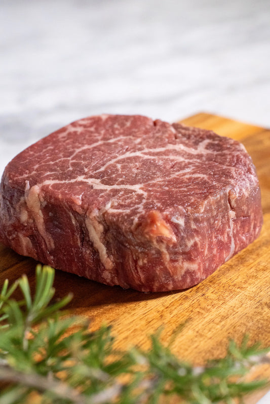 Raw Halal Wagyu filet steak on a wooden cutting board with rosemary on a a white granite top.

